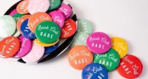 A stack of small colorful round pins that read "book club babe" in cursive script