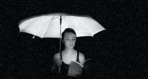grayscale photo of an Asian woman holding an umbrella and reading while it rains