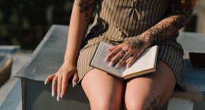 beige-skinned tattoed woman wearing a skirt and holding a book open on her lap
