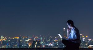light-skinned Asian business man sitting on a rooftop in a big city with light from a laptop shining on his face