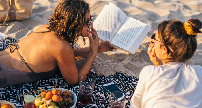 a photo of two women laying on the beach. One is reading a book